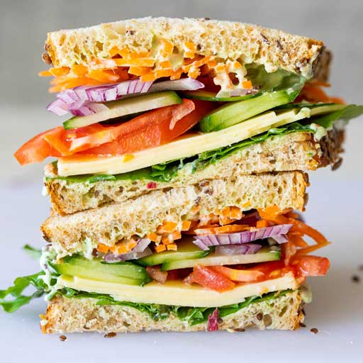 A stacked sandwich with cheese, red onions, avocado, tomato, meat, and spinach.